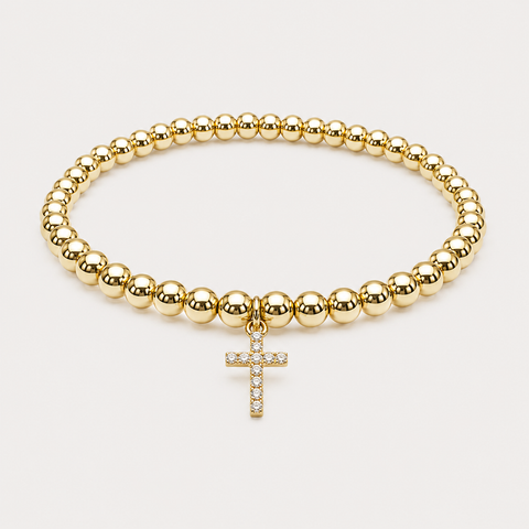 A gold beaded bracelet with a small cross charm adorned with clear stones, displayed on a light beige background.