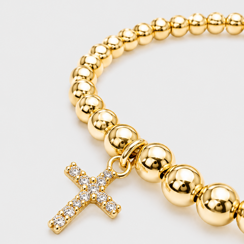 A gold beaded bracelet with a small gold cross charm encrusted with clear crystals, shown on a light gray background.