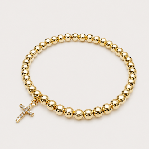 A gold beaded bracelet with a small cross charm, photographed on a clean, light beige background.