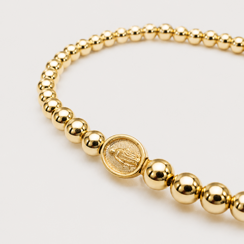 A gold beaded bracelet featuring a central round medallion with an embossed religious figure, photographed on a clean light gray background.