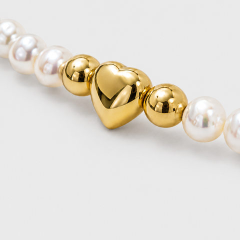 A close-up of a beaded strand featuring glossy white pearl-like round beads and polished gold metallic heart and sphere accents on a light gray background.