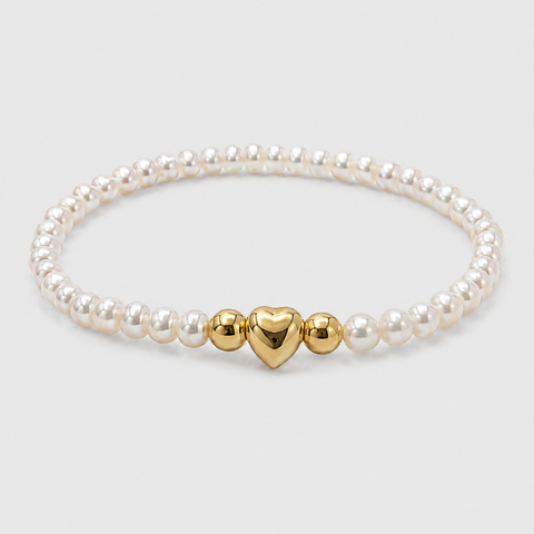 A pearl stretch bracelet featuring a central gold heart charm flanked by two gold ball charms, displayed on a light gray background.