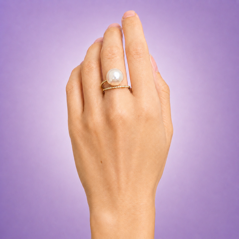 Model wearing 14K yellow gold pavé diamond double band white pearl ring on middle finger hand lifestyle shot purple background