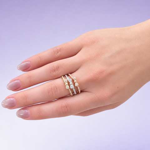 Yellow, white, and rose gold pavé diamond bow rings stacked on a finger against a lavender background.