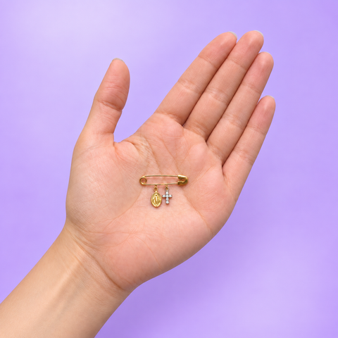 A hand holding a solid 18K yellow gold safety pin featuring a Miraculous Medal charm and a diamond cross charm, shown against a soft lilac background to demonstrate the brooch's true size and scale.