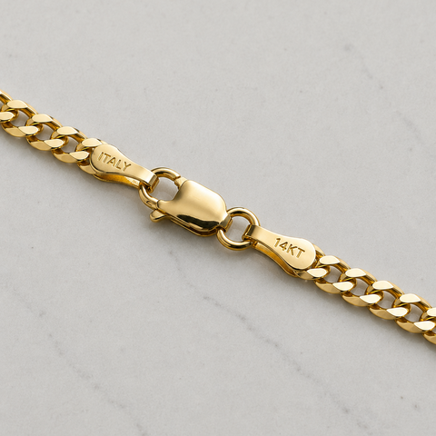 A close-up of a gold chain necklace showing the lobster clasp and engraved details, including 'ITALY' and '14KT', on a light gray marble surface.