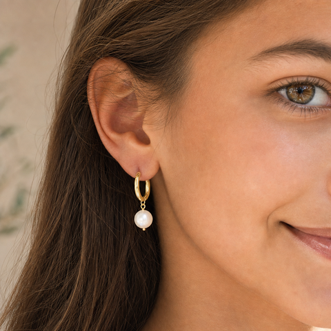 A close-up of a woman wearing a delicate gold huggie hoop earring with a dangling white pearl.