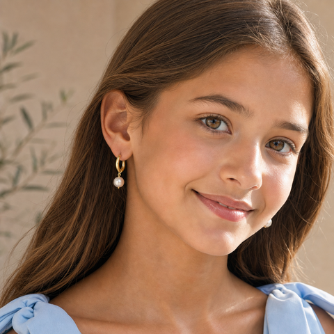 A young girl with long brown hair wearing a light blue bow-shoulder top and a small gold hoop earring with a white pearl drop, shown in a close-up portrait.