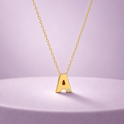 A gold initial necklace featuring the letter 'A' pendant with a small clear gemstone inset, displayed on a lavender pedestal against a soft lavender background.