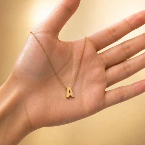 A gold initial necklace with the letter 'A' pendant, displayed on an open hand against a warm, soft-focus beige background.