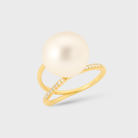 A gold ring featuring a large, round, creamy off-white pearl on a slender, openwork gold band accented with small clear stones, set against a light gray background.