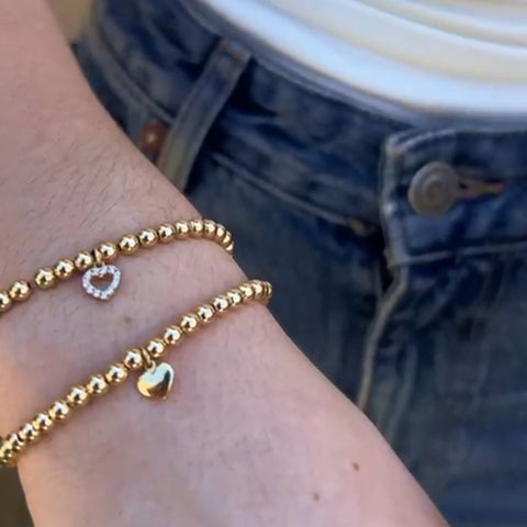 14K yellow gold heart charm bead bracelet worn on woman's wrist layered with second gold beaded bracelet, lifestyle stacking view showing how to layer
