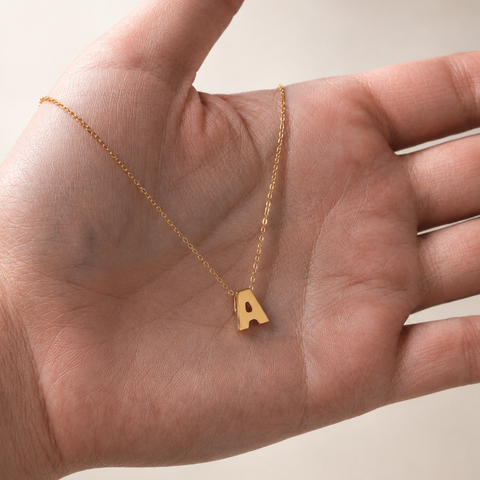 A delicate gold chain necklace with a small capital letter 'A' pendant, resting on an open hand against a soft beige background.