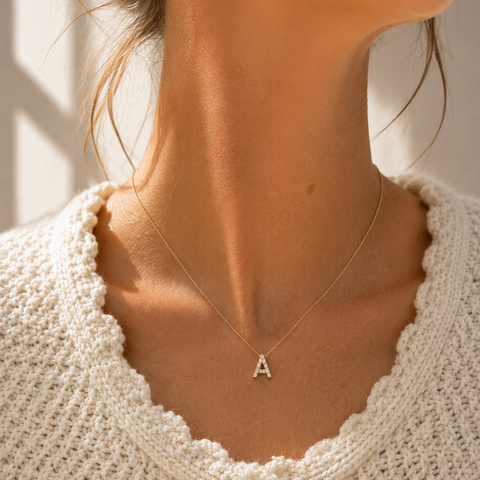 A close-up lifestyle shot of a model wearing a delicate gold necklace with a single letter 'A' pendant, styled with a cream-colored textured knit cardigan.