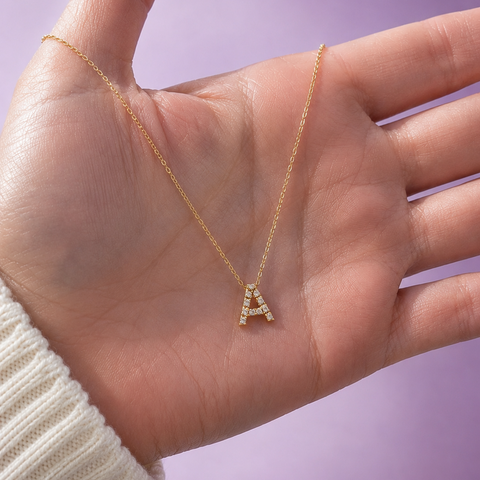 A gold initial necklace with the letter 'A' adorned with small diamonds, displayed on an open hand to show proportion against a soft purple background.