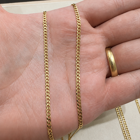 A close-up of a person's hand displaying two gold chain necklaces, with one chain draped over the back of the hand and the other hanging down. A gold ring is also visible on the finger.