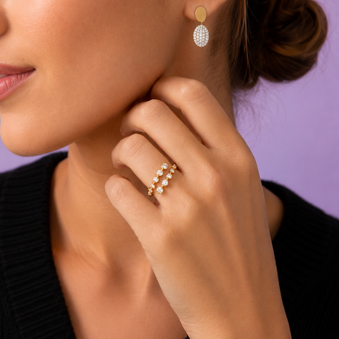 A close-up lifestyle shot of a woman wearing a delicate gold ring with small clear stones on her finger, and a matching gold drop earring with a textured white stone cluster. She is wearing a black ribbed sweater against a solid lavender-purple background.