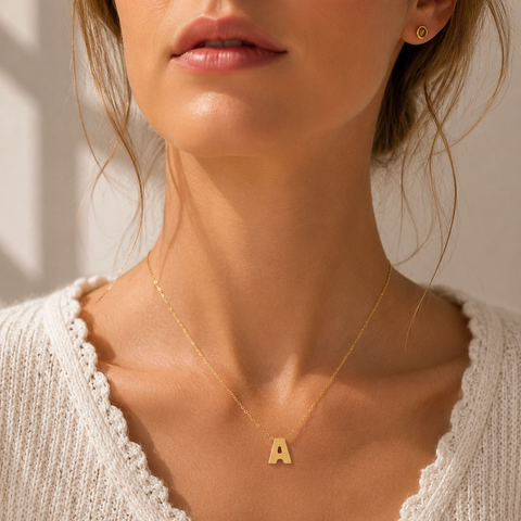 A close-up of a woman wearing a delicate gold necklace with a small capital letter 'A' pendant, styled with a white knit cardigan.